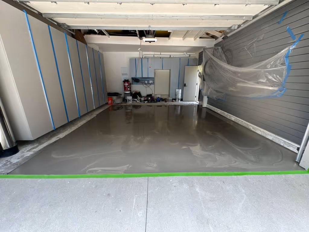 Garage interior with fresh, smooth, wet concrete floor being leveled, plastic-covered objects on the right wall.