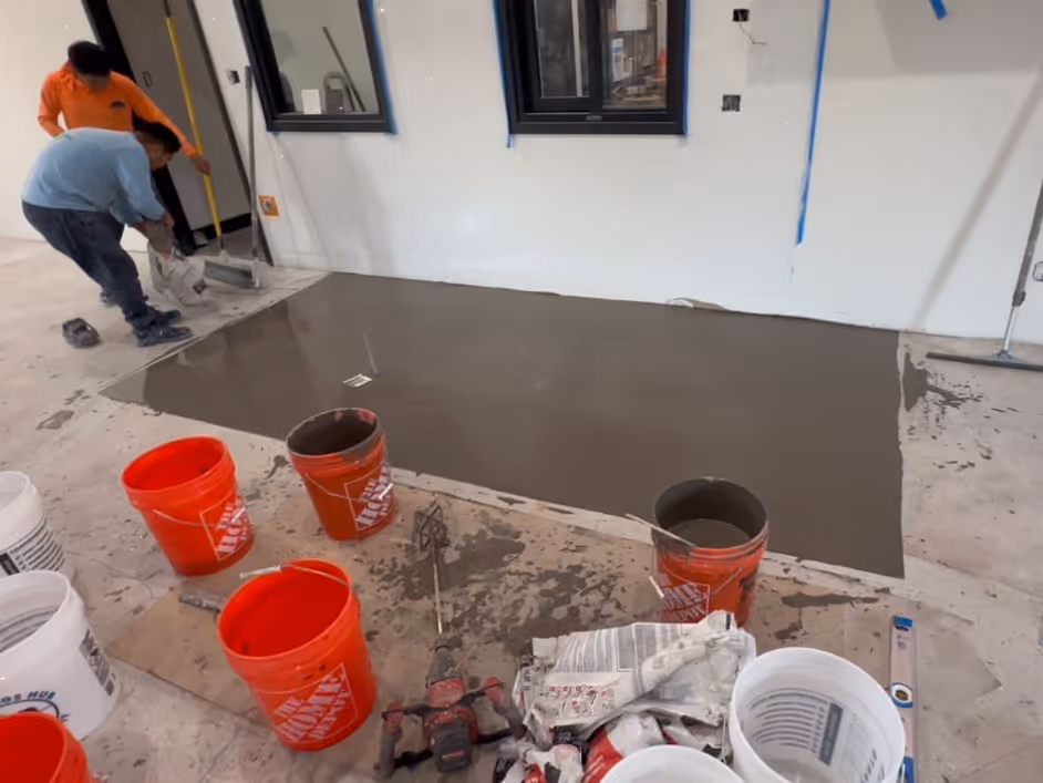 Two workers spreading self-leveling concrete on a floor inside a room with tools and orange buckets nearby.
