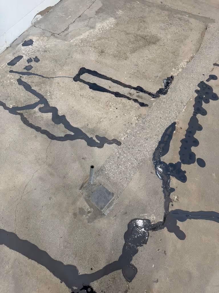Concrete floor with multiple cracks sealed with black crack filler around a metal pipe sticking out near a small square patch.