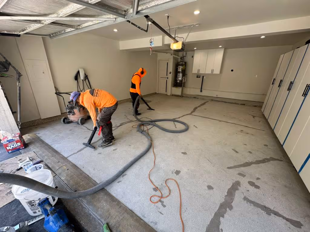 Two workers wearing orange shirts vacuuming and preparing a concrete garage floor.