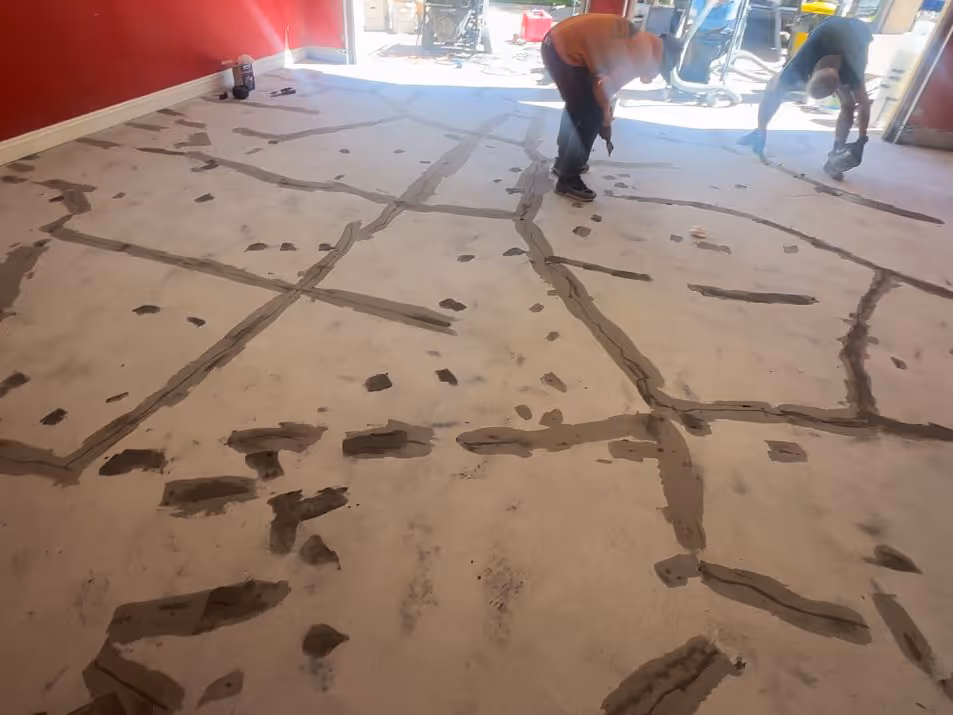 Two workers applying patching material to repair cracks on an indoor concrete floor with red walls.