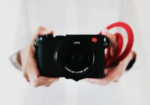 Closeup of photographer's hands holding a Leica camera with a red strap on a blurred white background