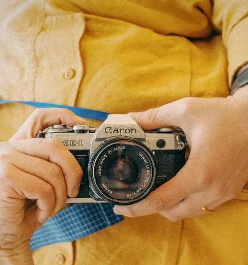 Photographer's hands and chest wearing a yellow shirt taking a selfie on a Canon AE-1 camera with a blue strap