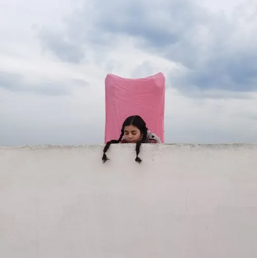 Girl with braids hanging over the edge of a white concrete wall with pink cloth behind her by Omar Eltahan
