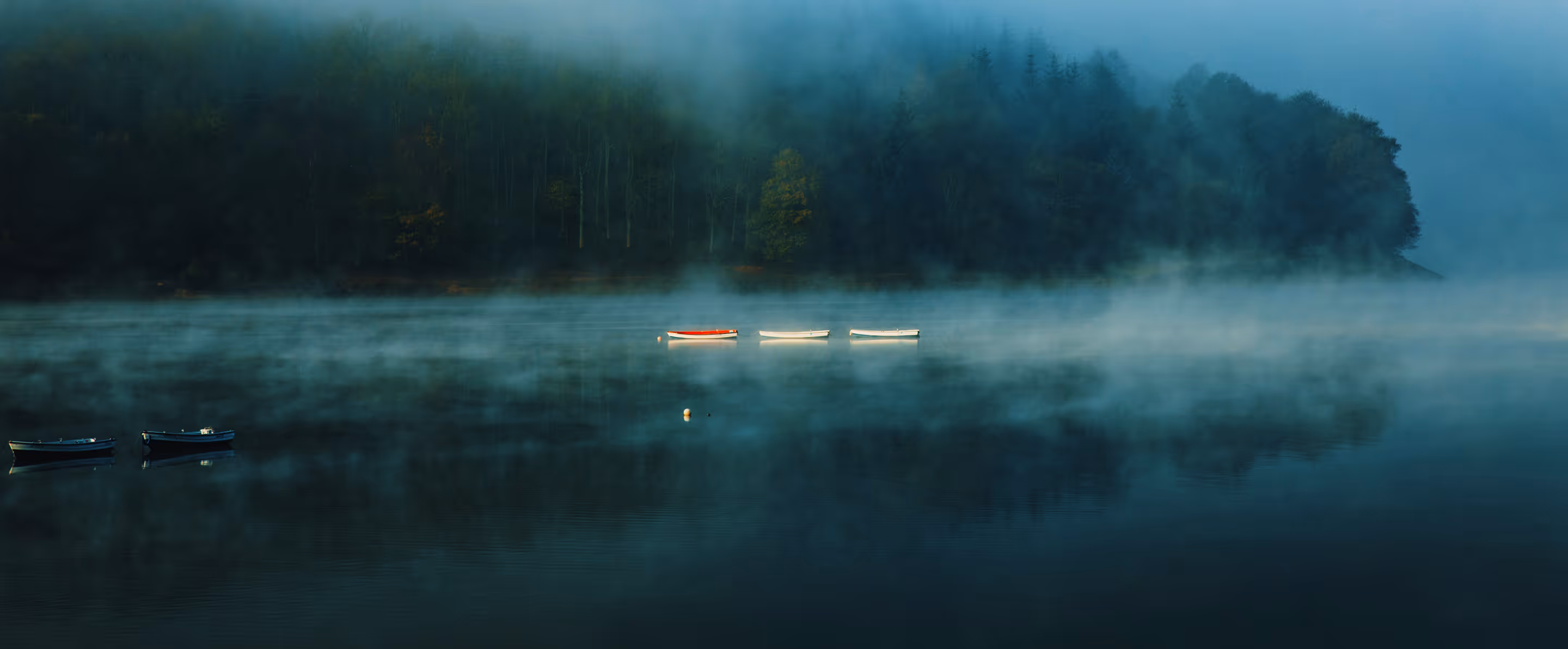 Five small boats floating on a misty lake with a forested shore in the background.
