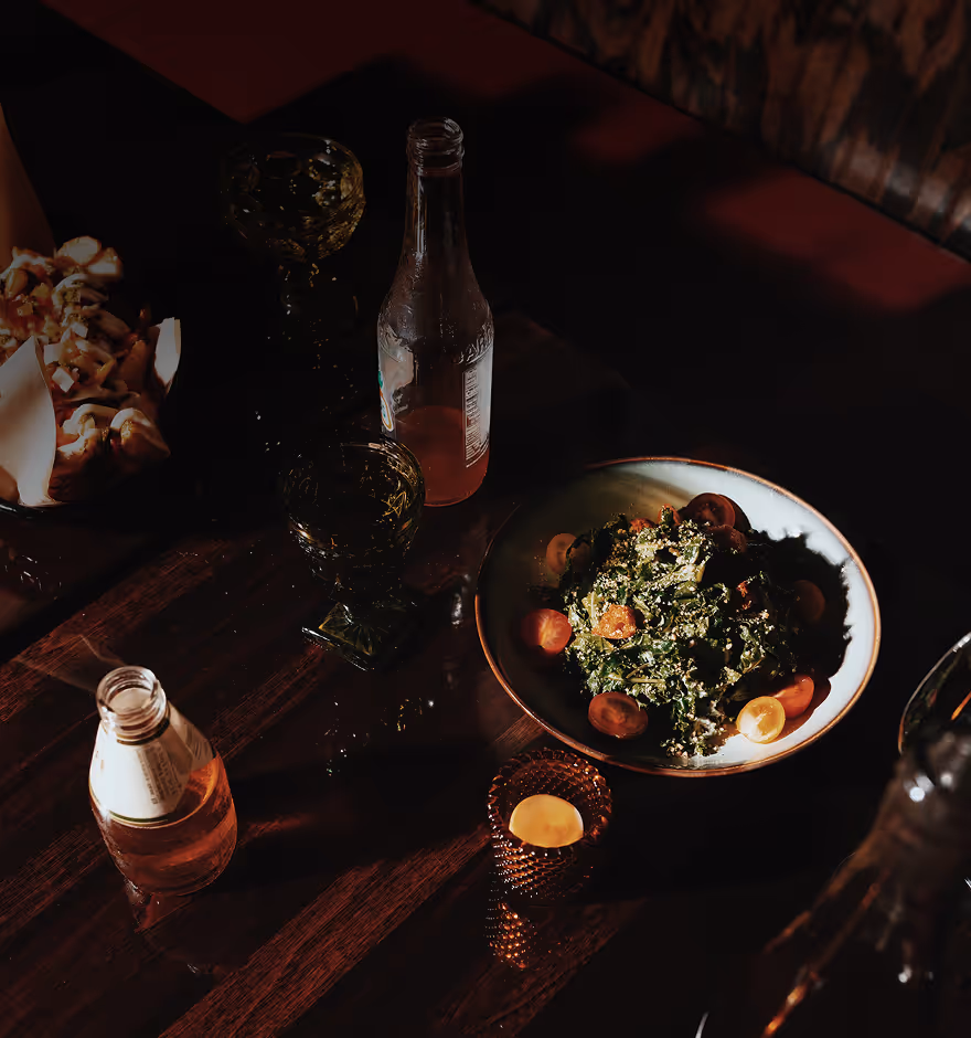 Film filter on a dark wooden table with a bowl of salad garnished with cherry tomatoes, two glass bottles, two vintage glass cups, a basket of popcorn, and a lit candle in a decorative holder.