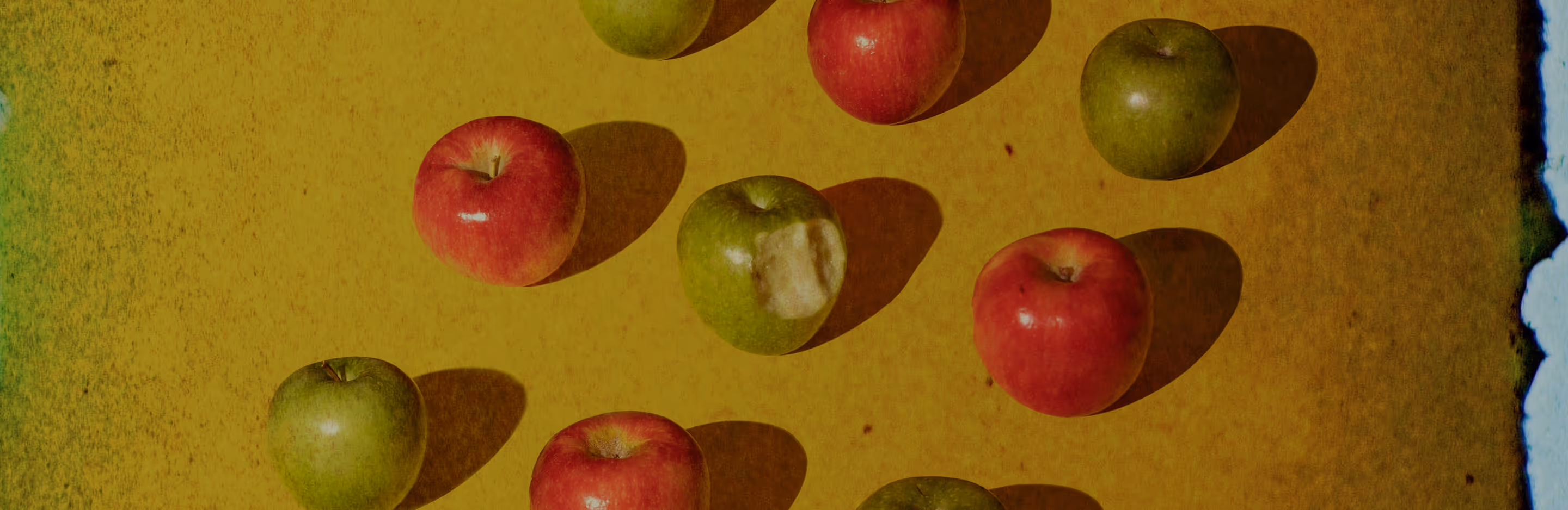 Film burn overlay added to image of red and green apples, one with a bite, arranged on a textured yellow surface.