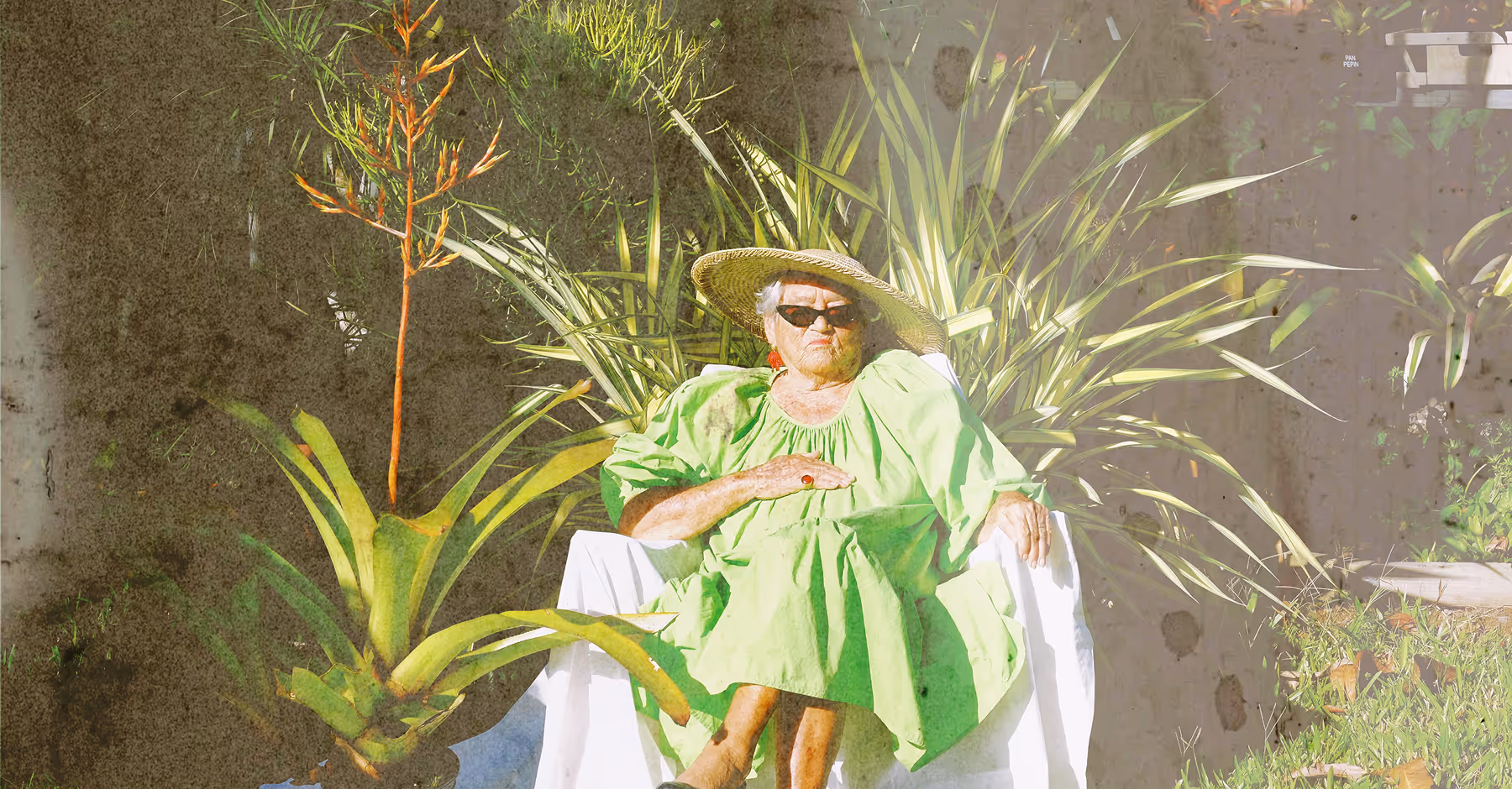 Expired film look overlaid on portrait of a woman wearing sunglasses, a wide-brimmed hat, and a bright green dress sitting on a white chair surrounded by large leafy plants.