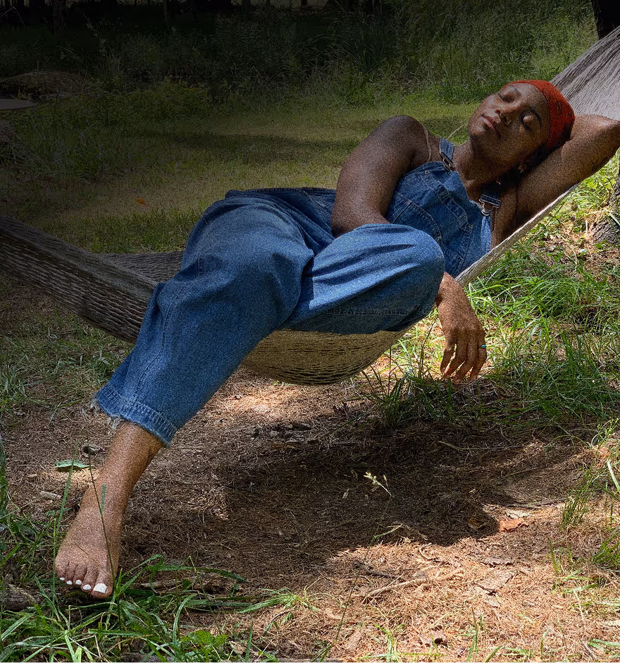 Grainy texture added on a photo of a person wearing denim overalls and a red headscarf relaxing on a hammock outdoors with eyes closed.