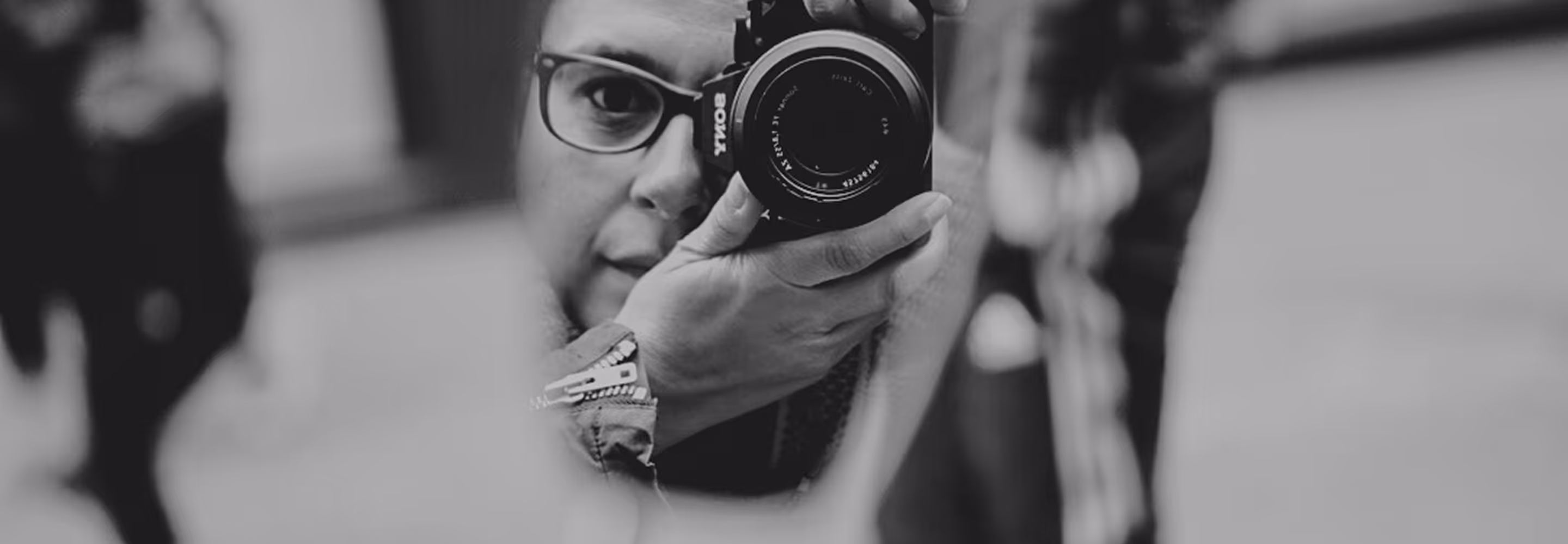 Close-up black and white photo of a photographer wearing glasses holding a Sony camera and taking a picture.