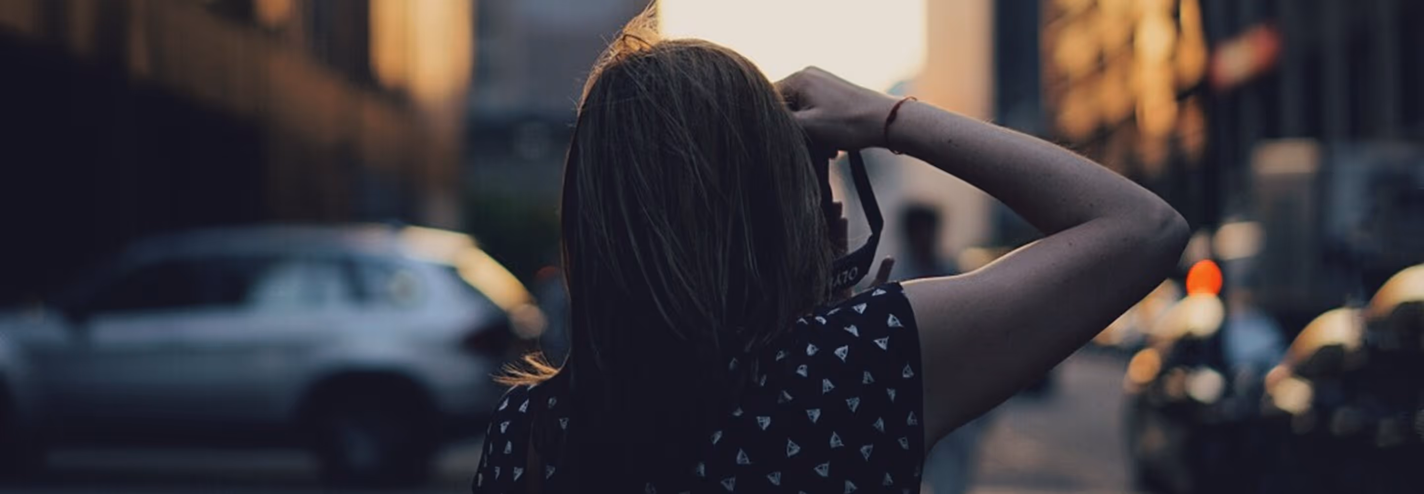 Person with long hair holding a camera up to their face on a city street during golden hour.