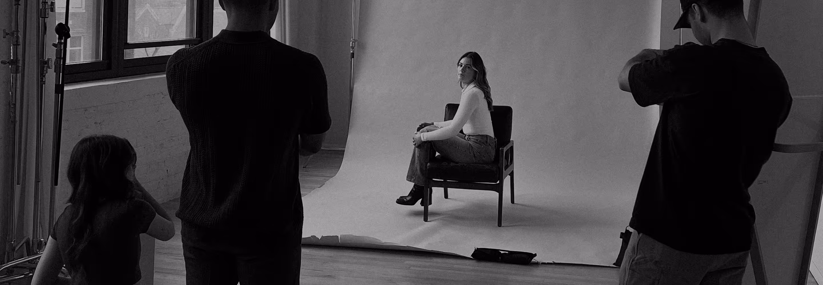 Photographers collaborating in a studio photoshoot with one person holding a camera and others watching model posing on chair.