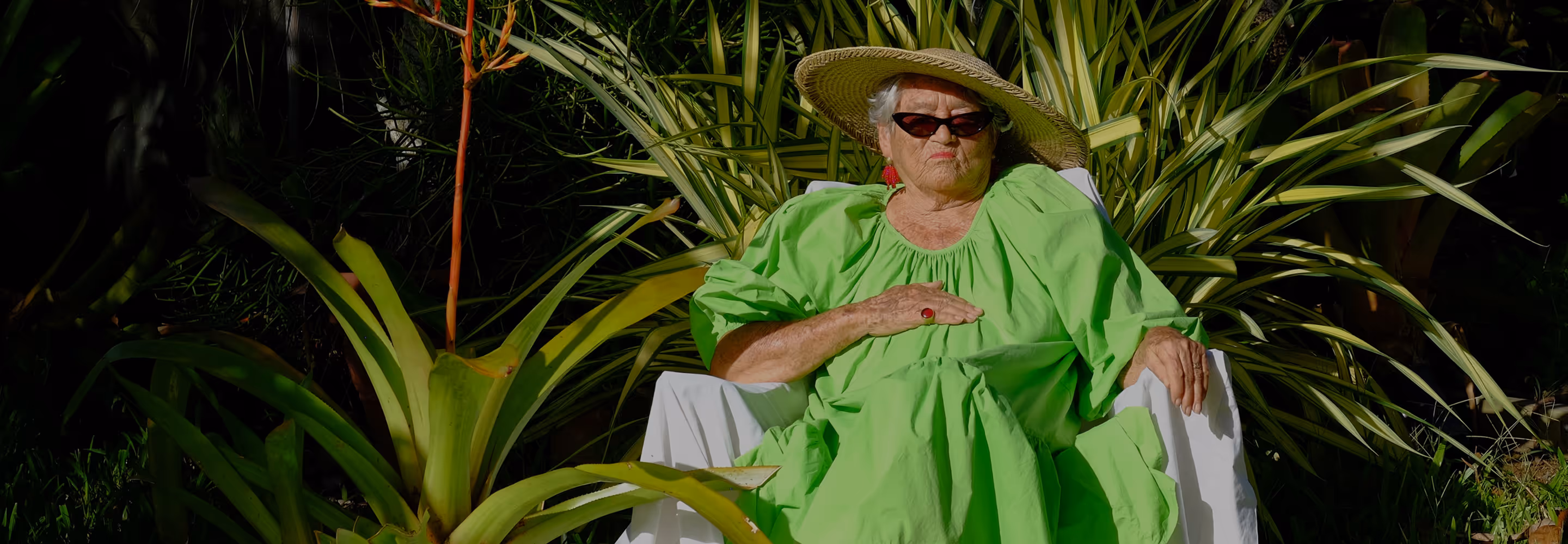 Older woman in bright green dress, wide-brimmed hat, and sunglasses sitting outdoors surrounded by green foliage.