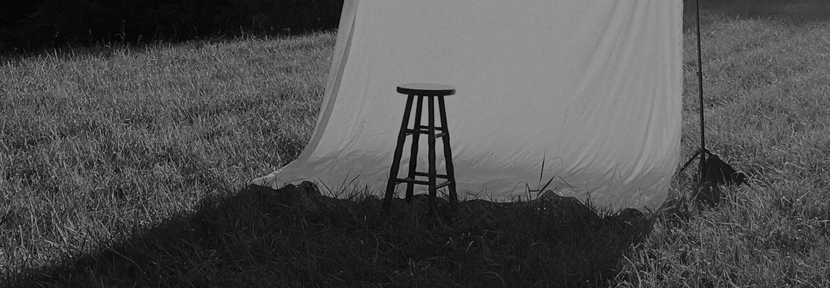 Wooden stool positioned on grass in front of a white backdrop held by stands in an outdoor photoshoot setting.