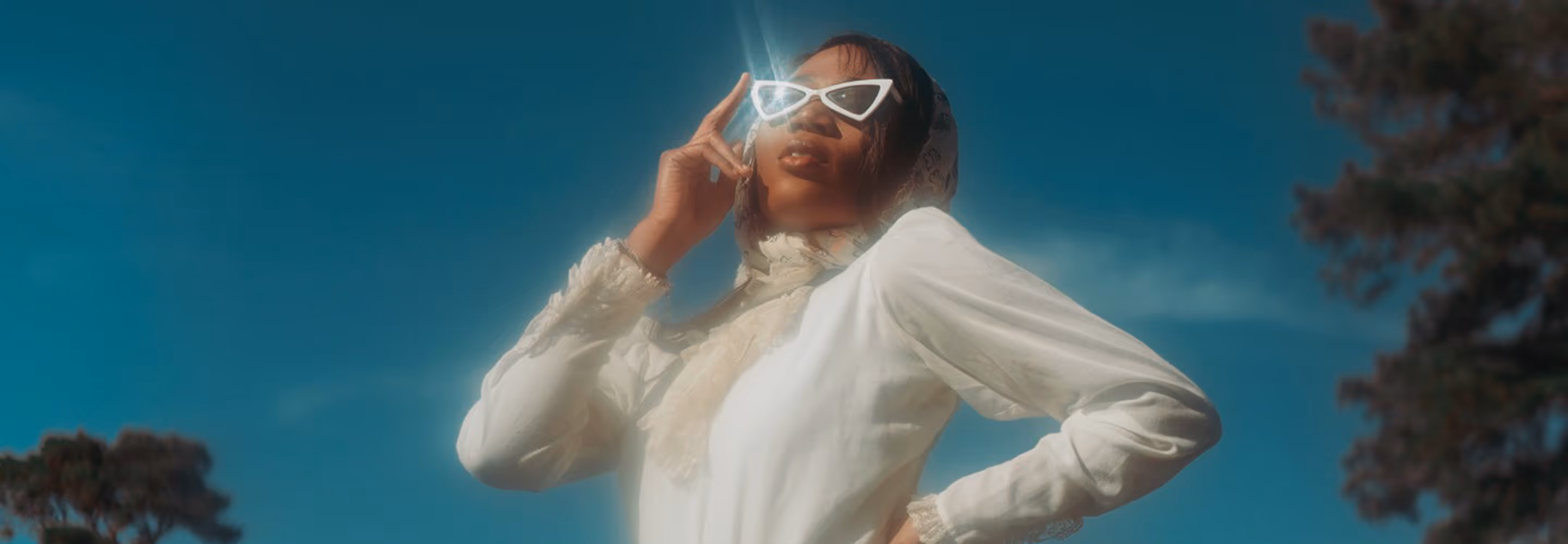Woman wearing white vintage-style dress and scarf with white cat-eye sunglasses under a clear blue sky.