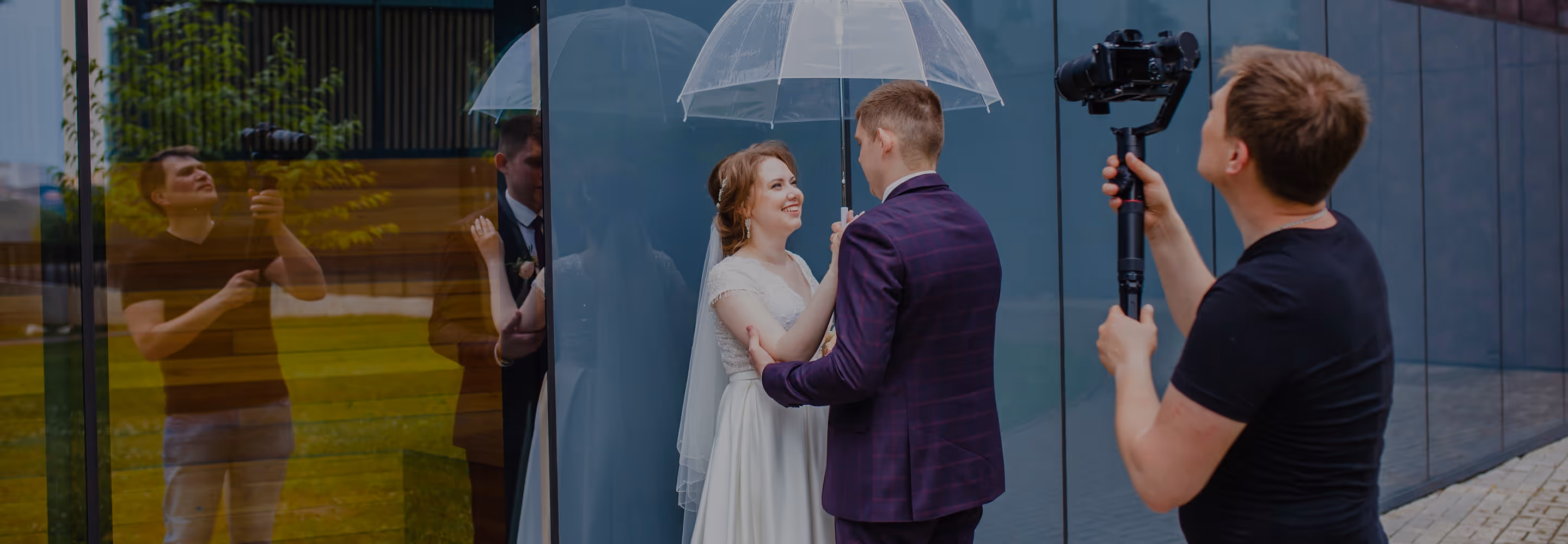 A wedding videographer films a smiling bride and groom under a transparent umbrella, standing by a reflective glass building.