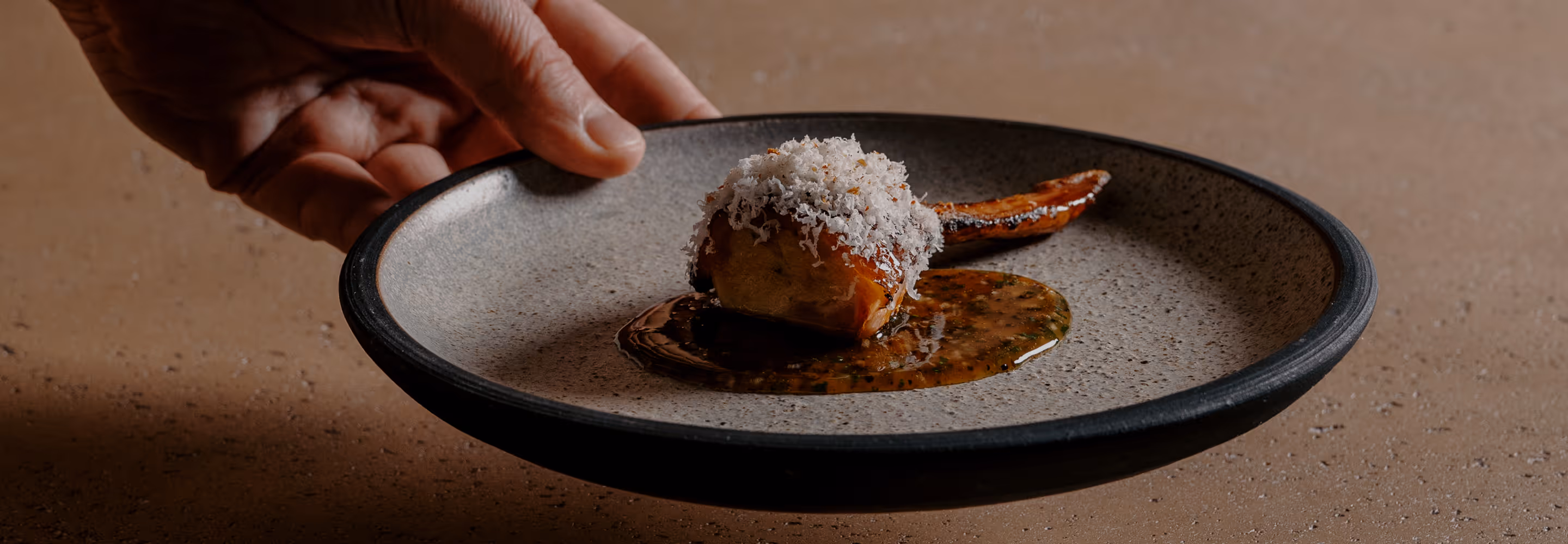 Hand holding a plate with a gourmet dish featuring a glazed piece of roasted vegetable topped with grated cheese and sauce.