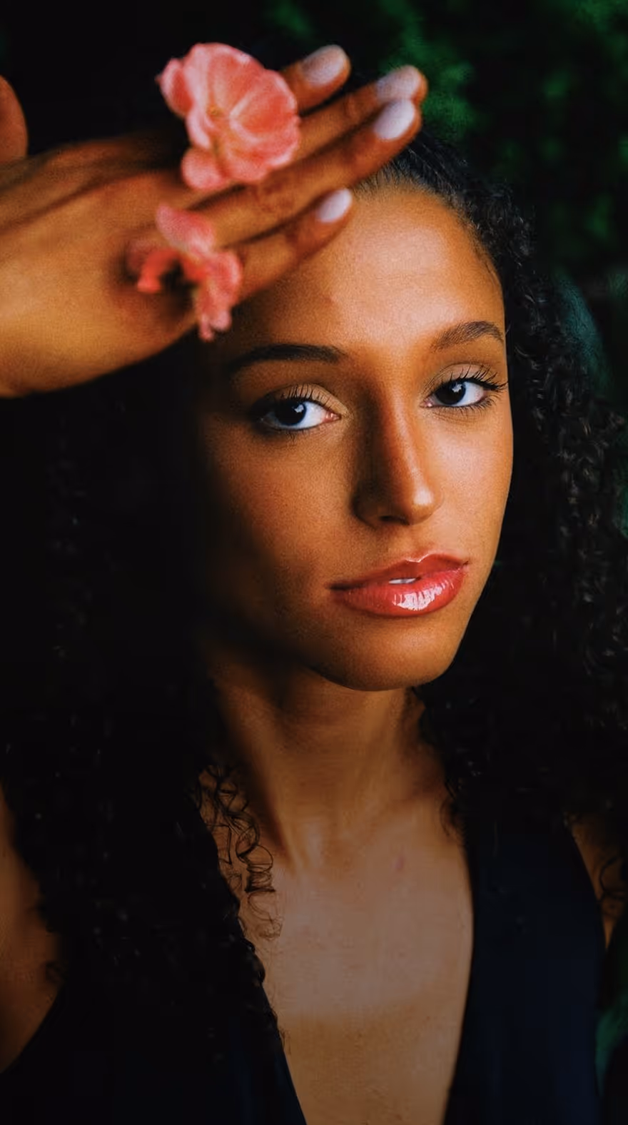 Close-up portrait of a woman holding pink flowers near her forehead, looking into the camera, taken by portrait photographer Tessa Burris.
