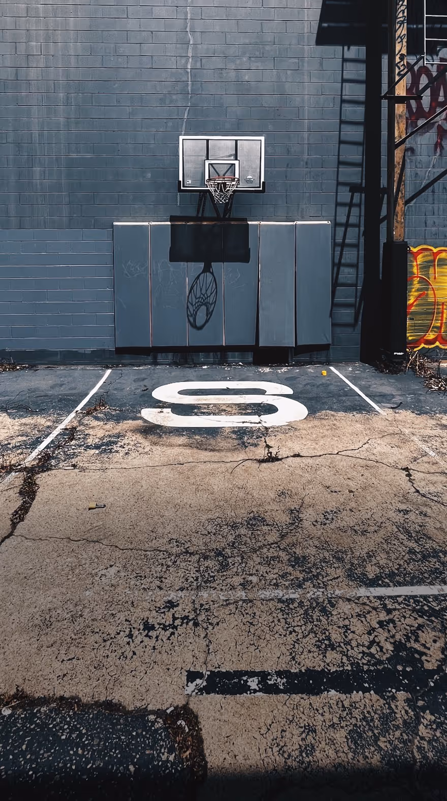 Outdoor basketball hoop mounted on a padded wall with cracked asphalt court and large white number 8 on the ground, taken by photographer Omeez Jones.