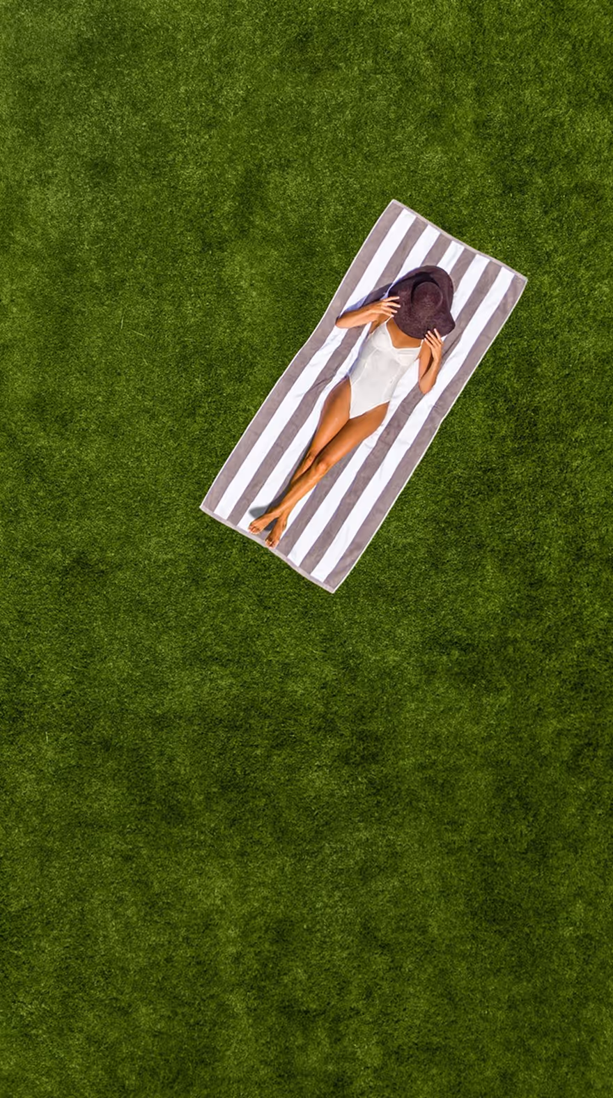 Woman in white swimsuit lying on striped towel on green grass, holding a large sunhat over her face, taken by commercial photographer Liza Gershman.