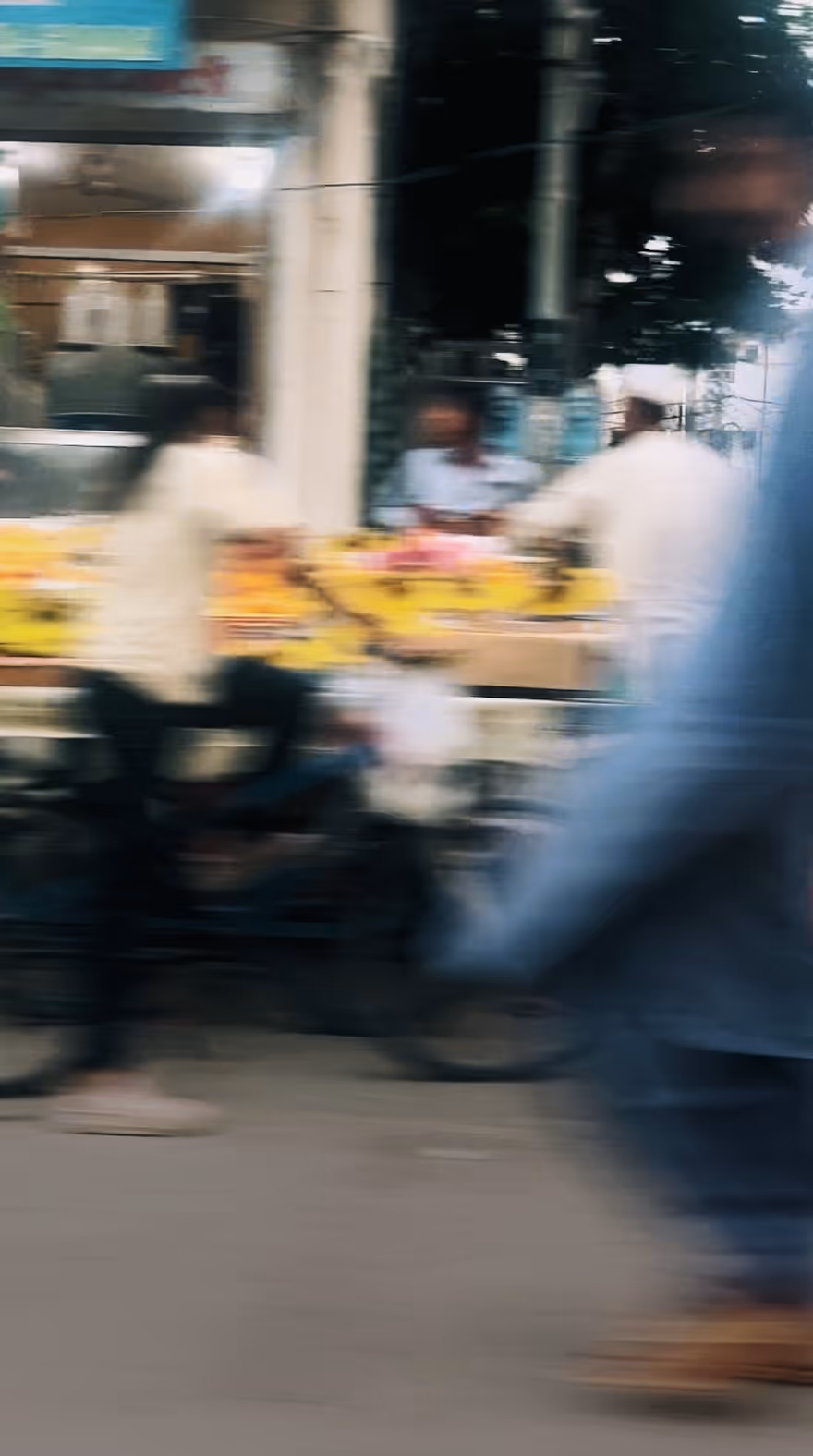 Blurred street scene with a person riding a bicycle and others near a fruit stall, taken by lifestyle photographer Simrah Farrukh.