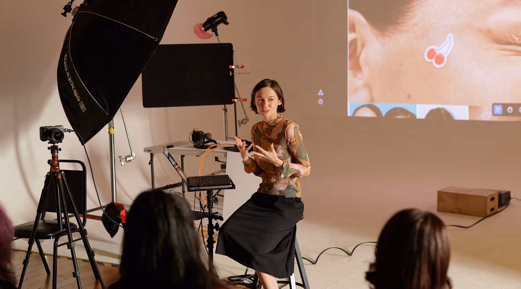 Woman sitting on a stool giving a presentation to an audience with a laptop, studio lighting, and a large projected image behind her.