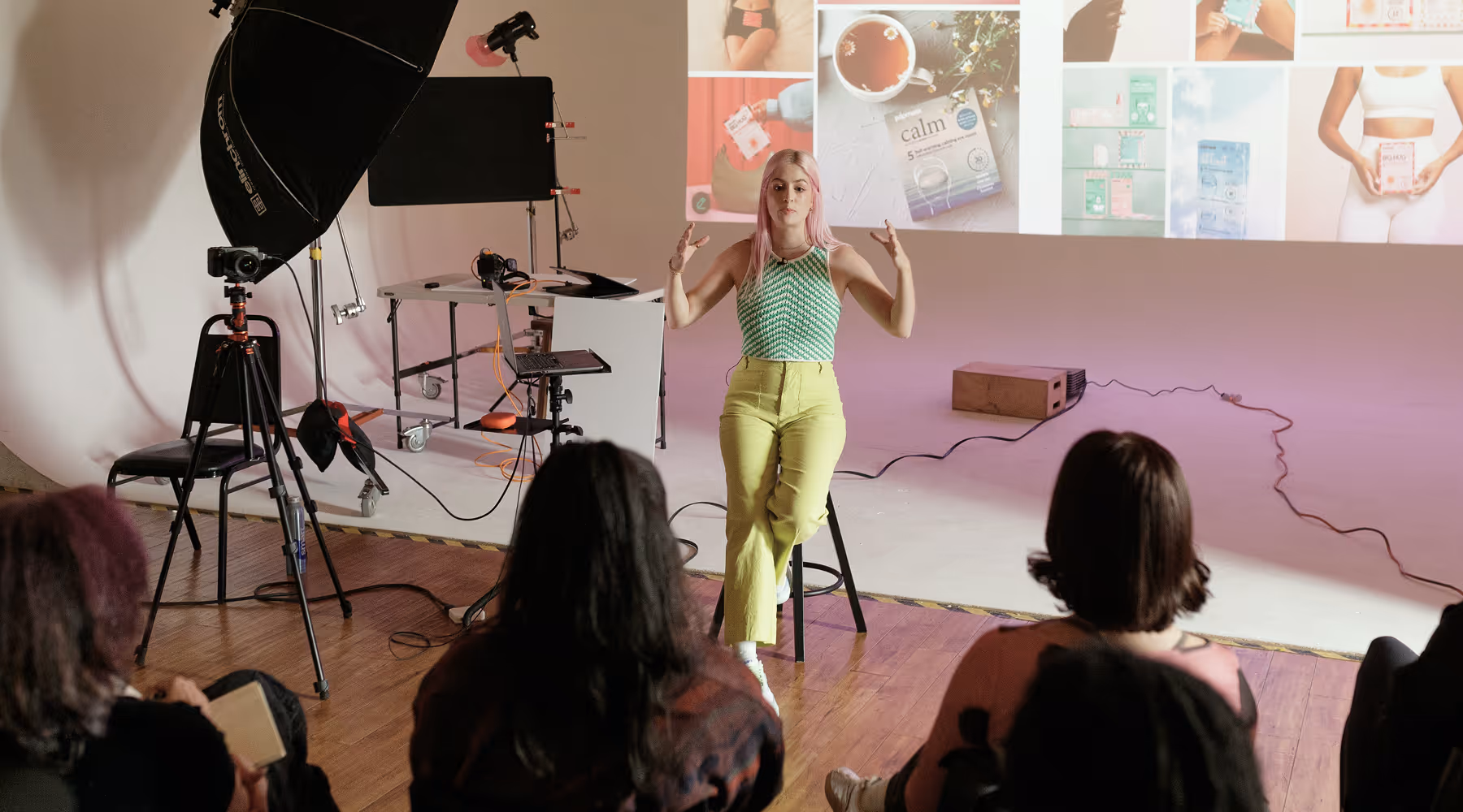 Woman with pink hair and green patterned top sitting on a stool speaking to an audience in a photography studio with projected images behind her.