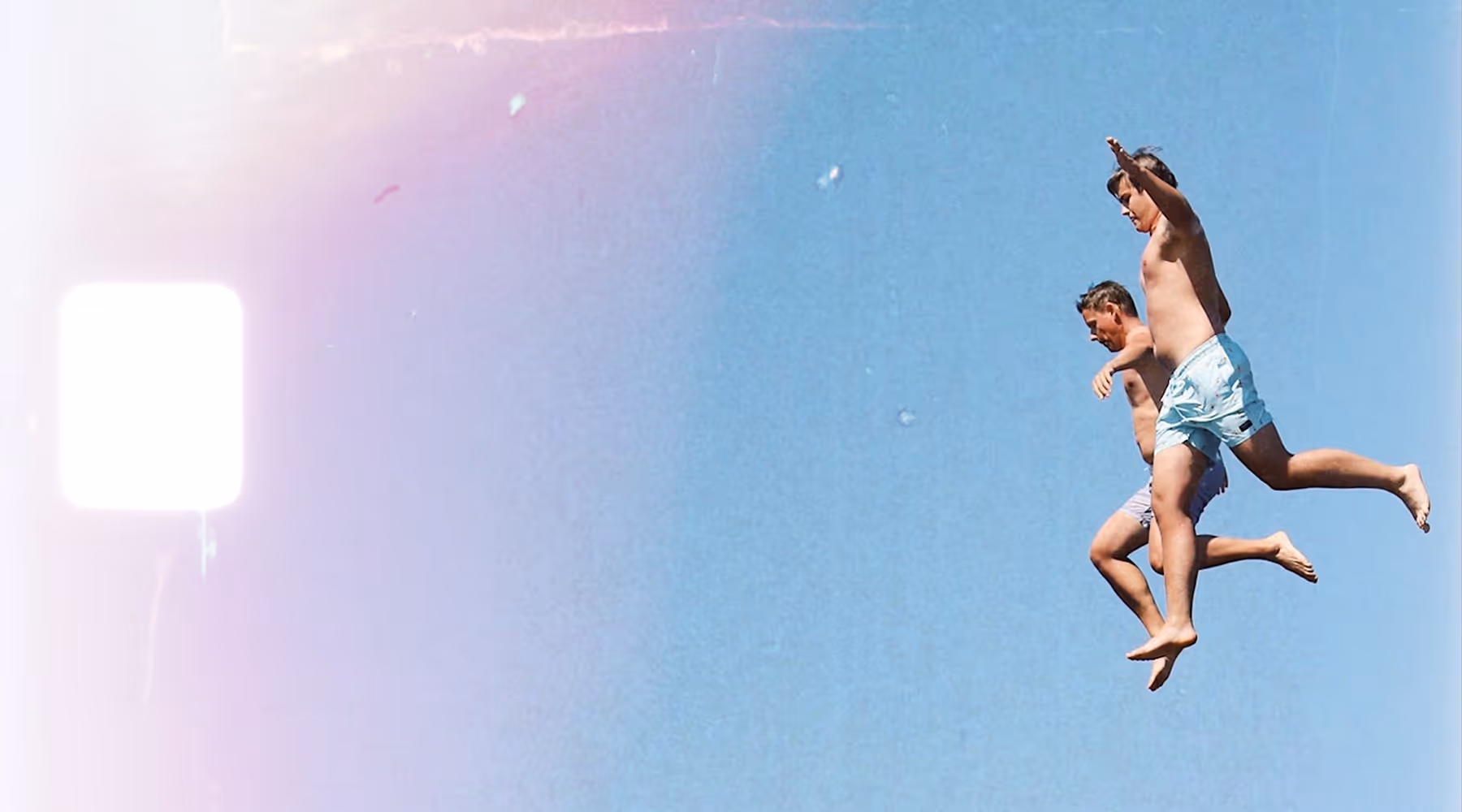 8mm film frame overlay on an image of two people in swim shorts jumping against a clear blue sky.