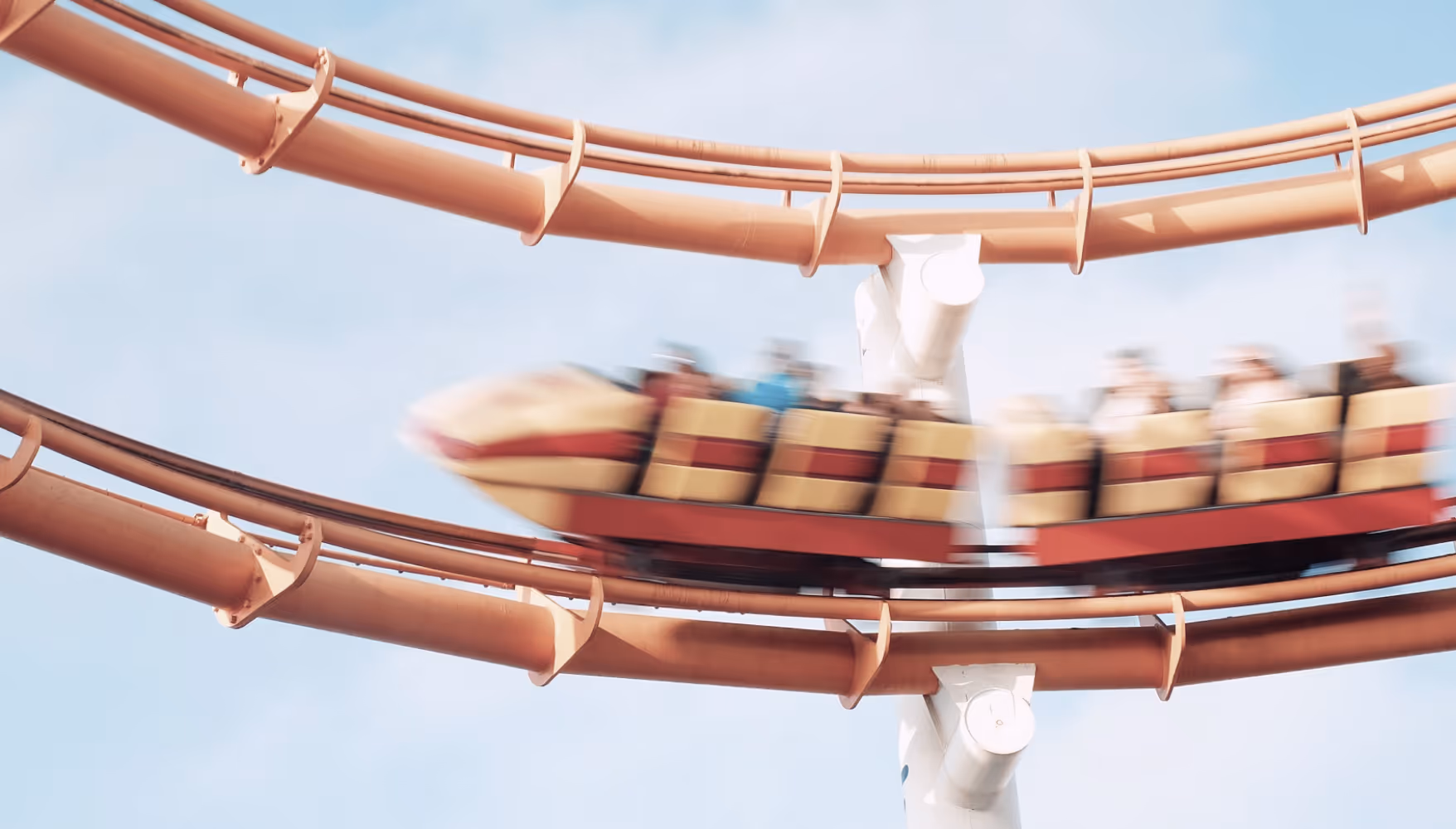 Muted image before color grading of a motion-blurred roller coaster train with riders speeding on red tracks against a clear blue sky.