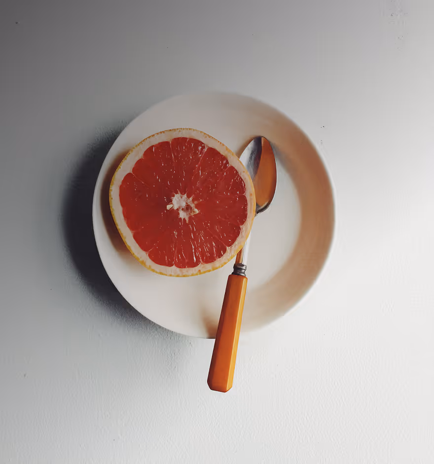 High contrast image of a half of a red grapefruit on a white plate with a spoon that has an orange handle.