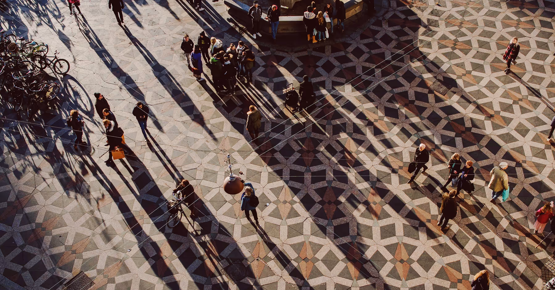 Overhead view of people walking and standing on a patterned plaza casting long shadows in sunlight edited for saturation