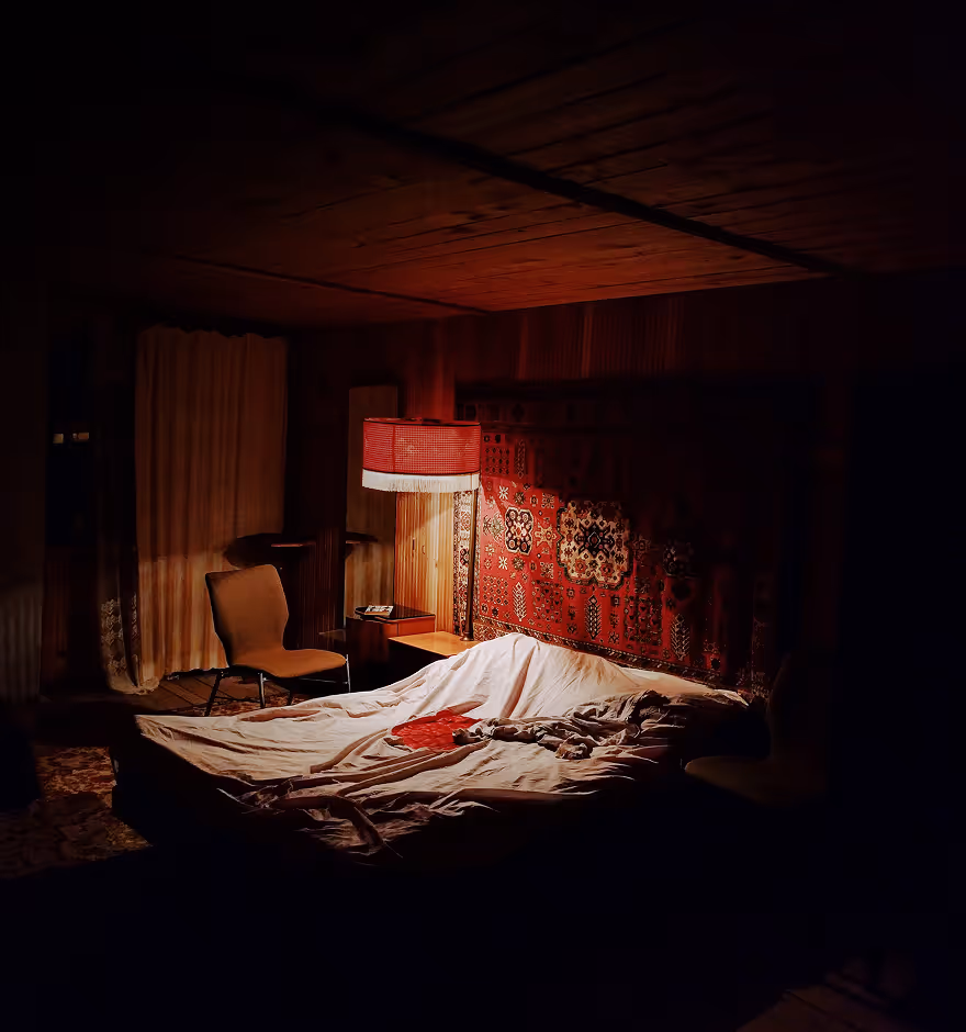 Dimly lit bedroom showing highlights and shadows with a bed covered in rumpled white sheets, a red patterned tapestry on the wall, a red lampshade glowing, and a chair beside the bed.