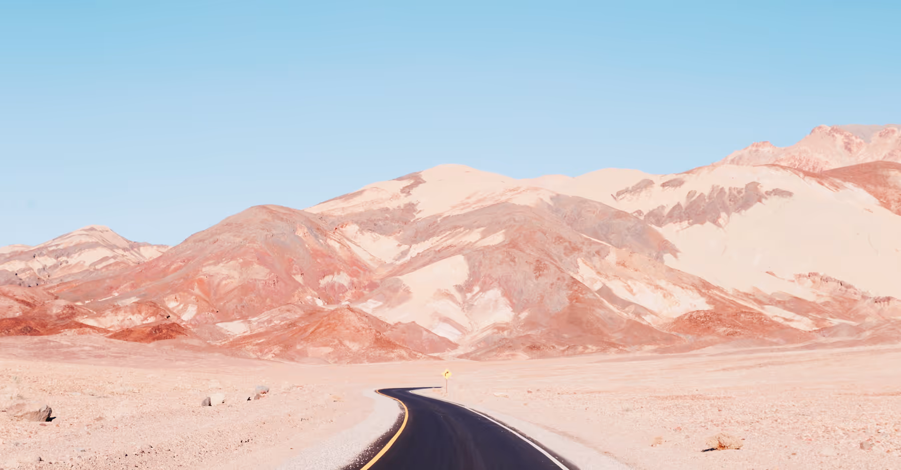 Saturated image of a curving road leading through a dry desert landscape with reddish hills under a clear blue sky.