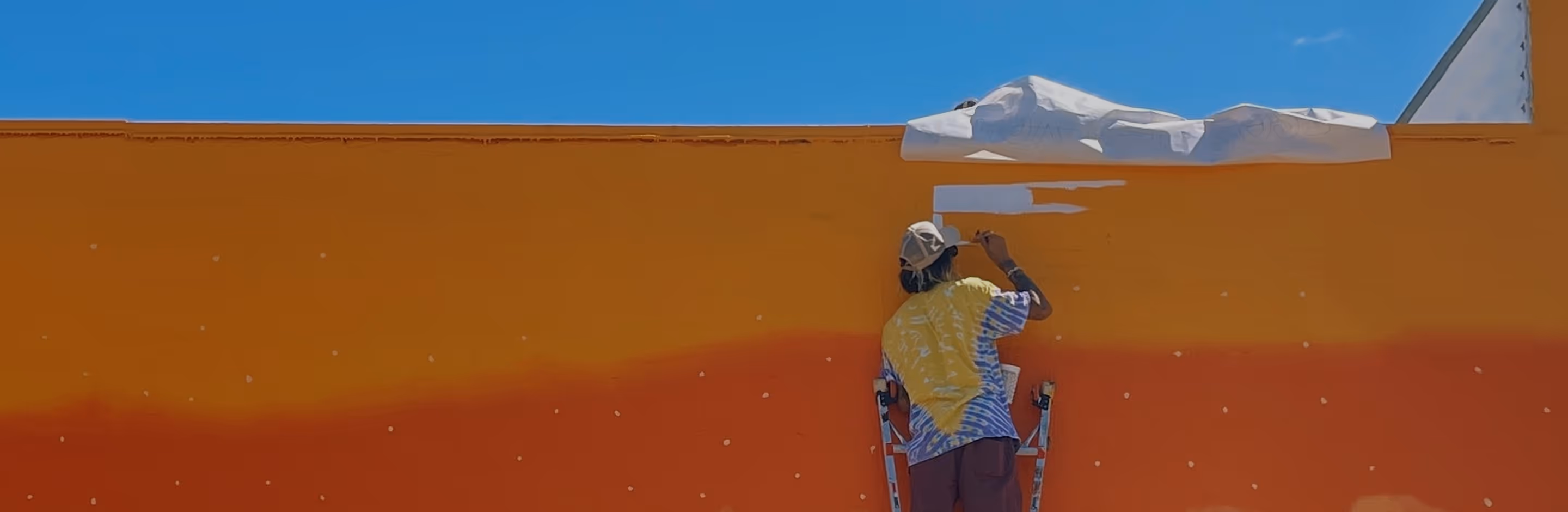 Colorful saturated photo of a person on a ladder painting the top section of a large orange and red mural wall under a clear blue sky.