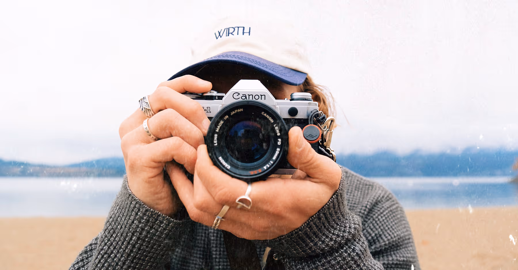 Distressed image of person wearing a cap and gray sweater holding a Canon film camera up to their face on a beach with blurred mountains in the background.