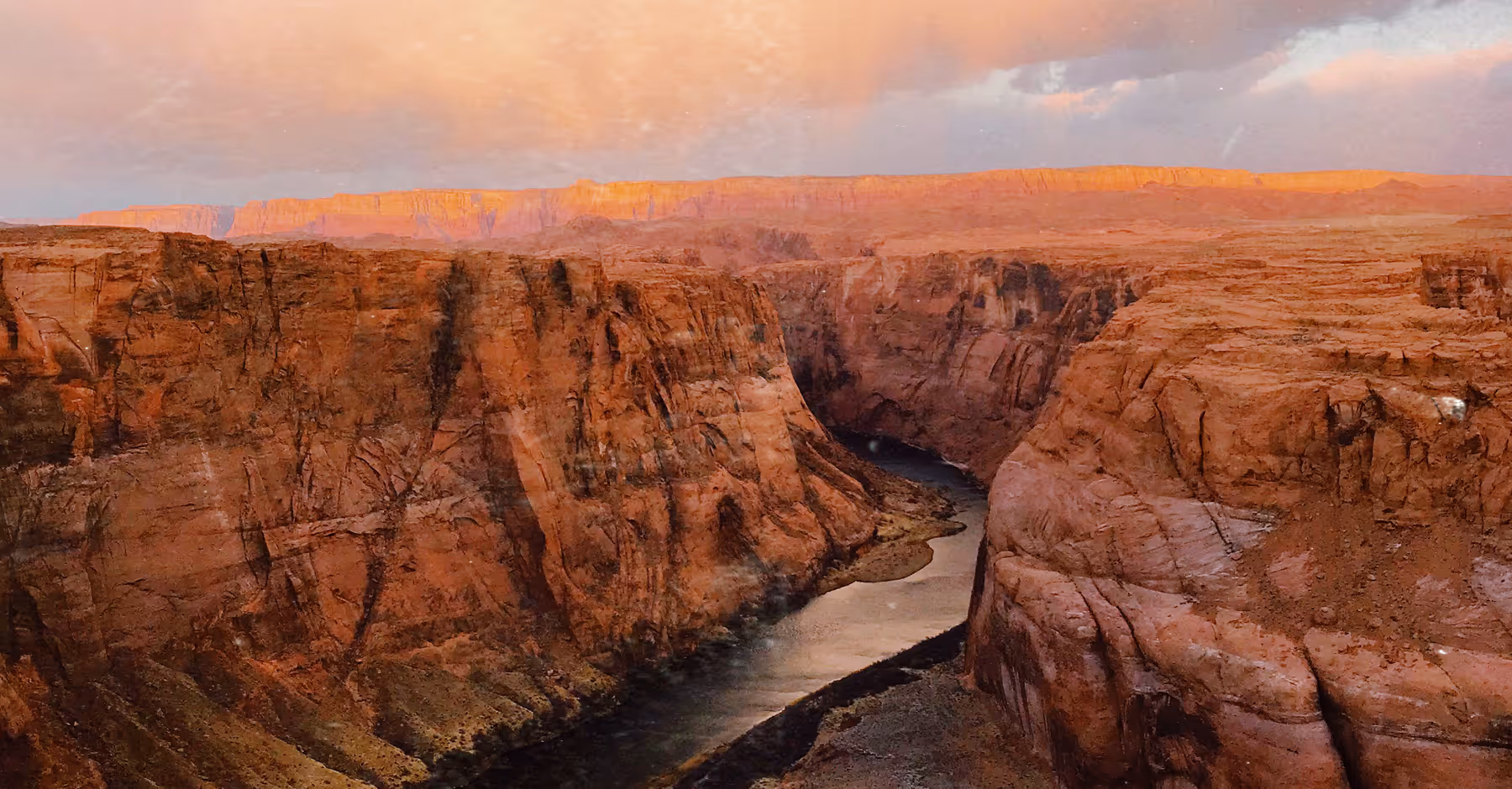 Wide view of a rocky canyon with a river flowing through it under a pastel-colored sky at sunset with a scratch texture overlay.
