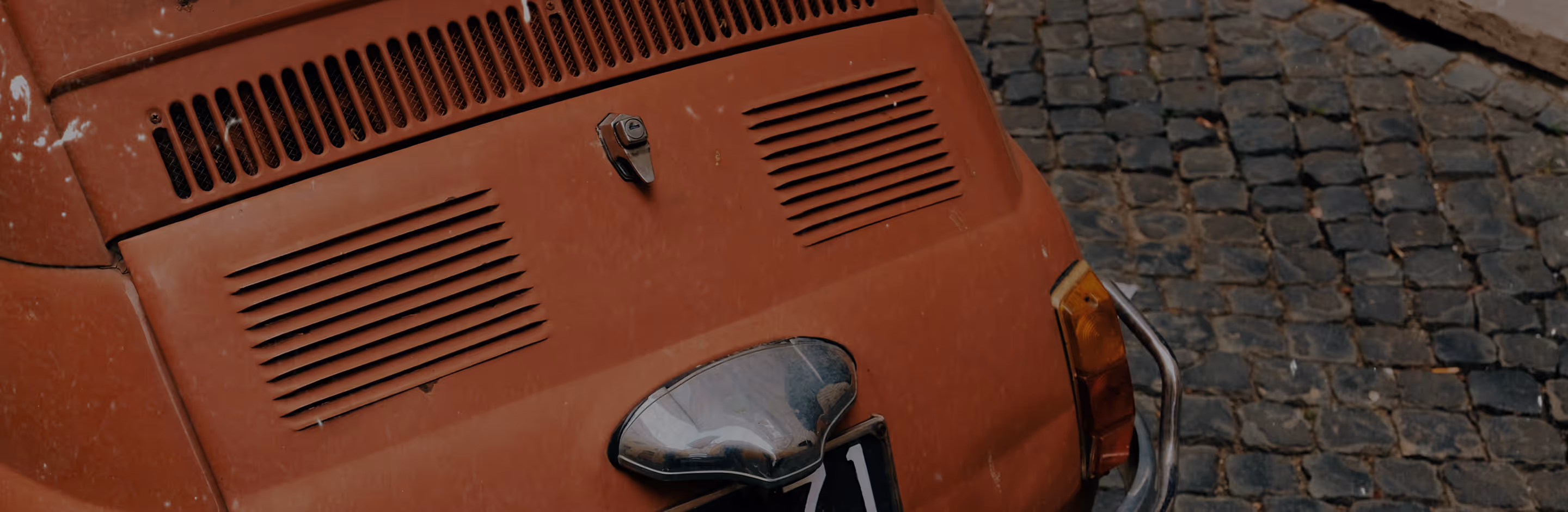 Scratched texture overlay on image of a rear view of a vintage orange car with air vents and a chrome handle parked on cobblestone pavement.