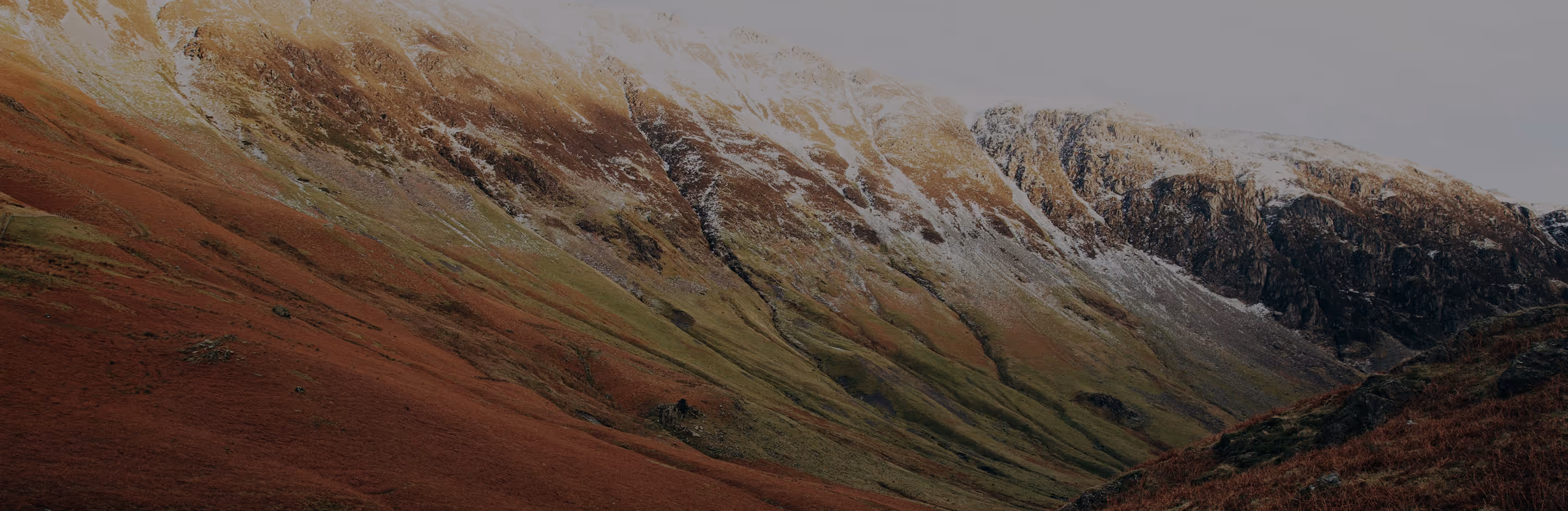 80s filter on a mountain slope with patches of snow, green grass, and red-brown vegetation under a light gray sky.