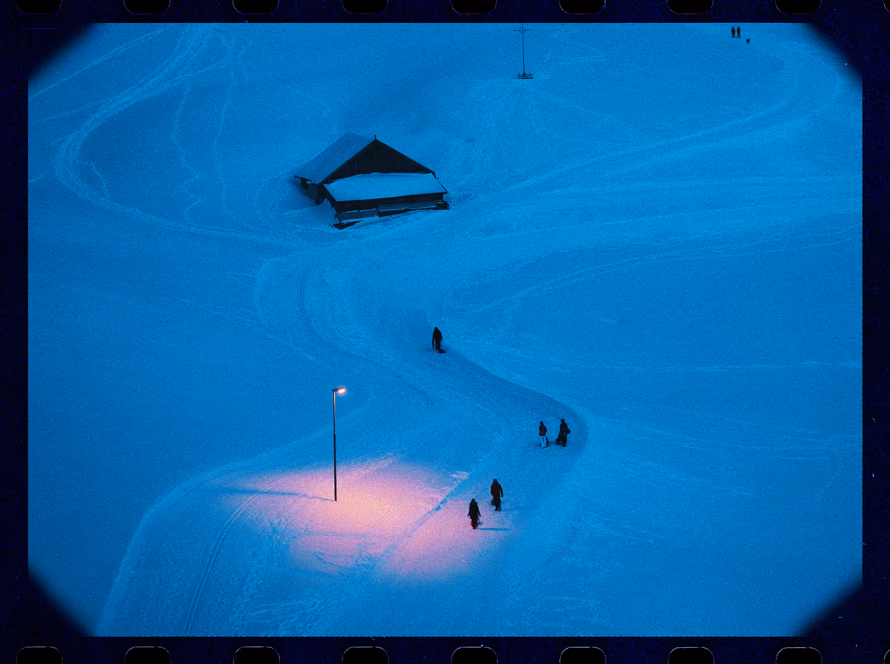 80s filter and film border on a snow-covered landscape with a small cabin, a lit streetlamp casting warm light, and several people walking along curved paths in the snow.