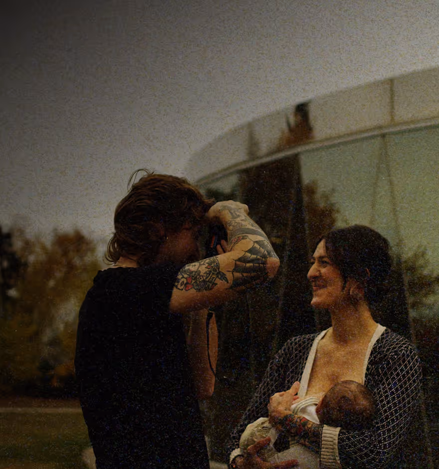 Film effect added to photo of tattooed photographer taking a photo of a smiling woman and baby outdoors near a curved glass building.