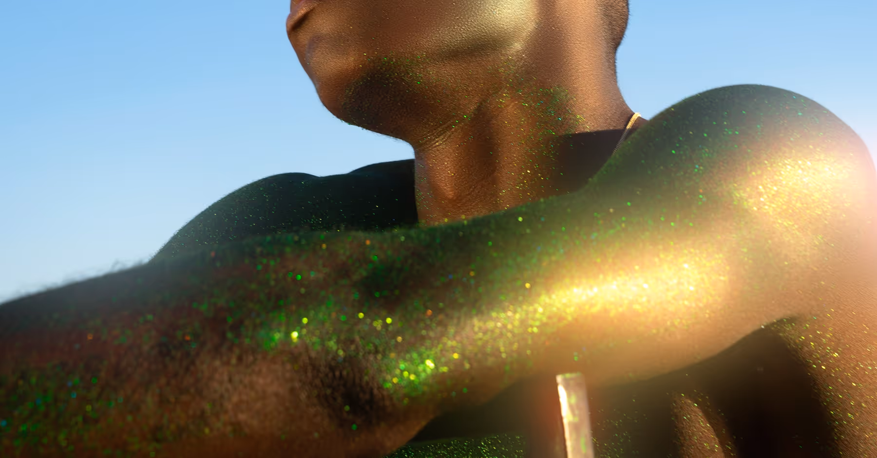Dreamy close-up of a person’s shoulder, neck, and arm covered in shimmering green and gold glitter against a clear blue sky.