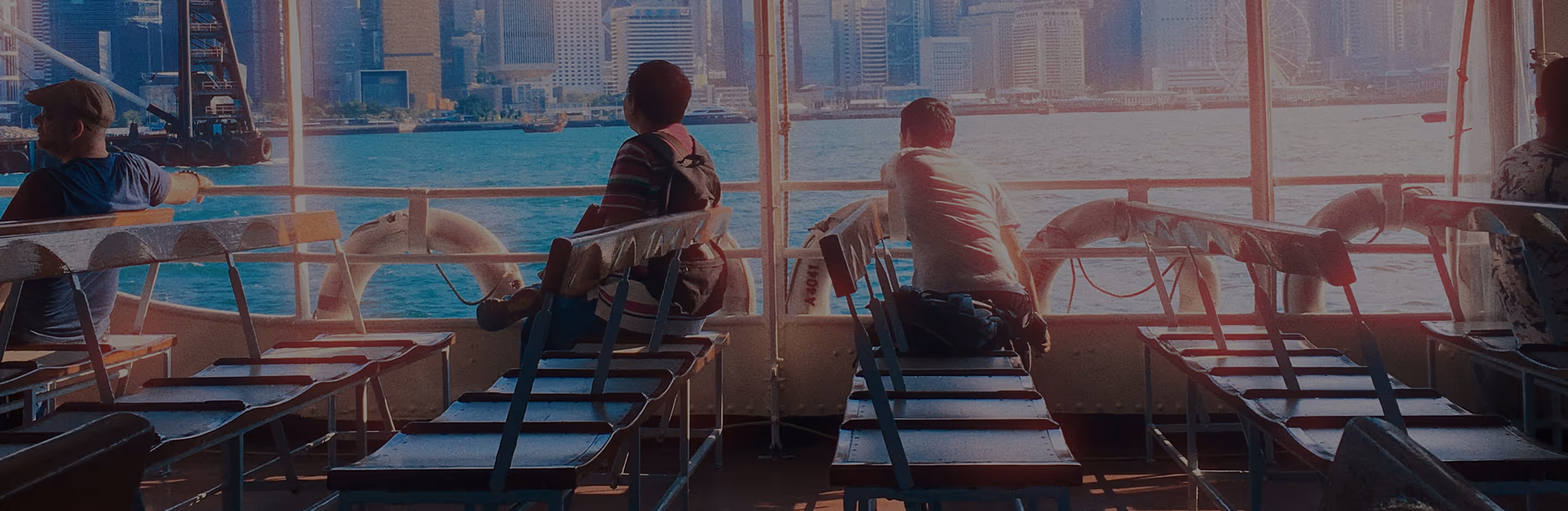Cinematic photo of people sitting on empty benches on a ferry with a city skyline across blue water in the background.