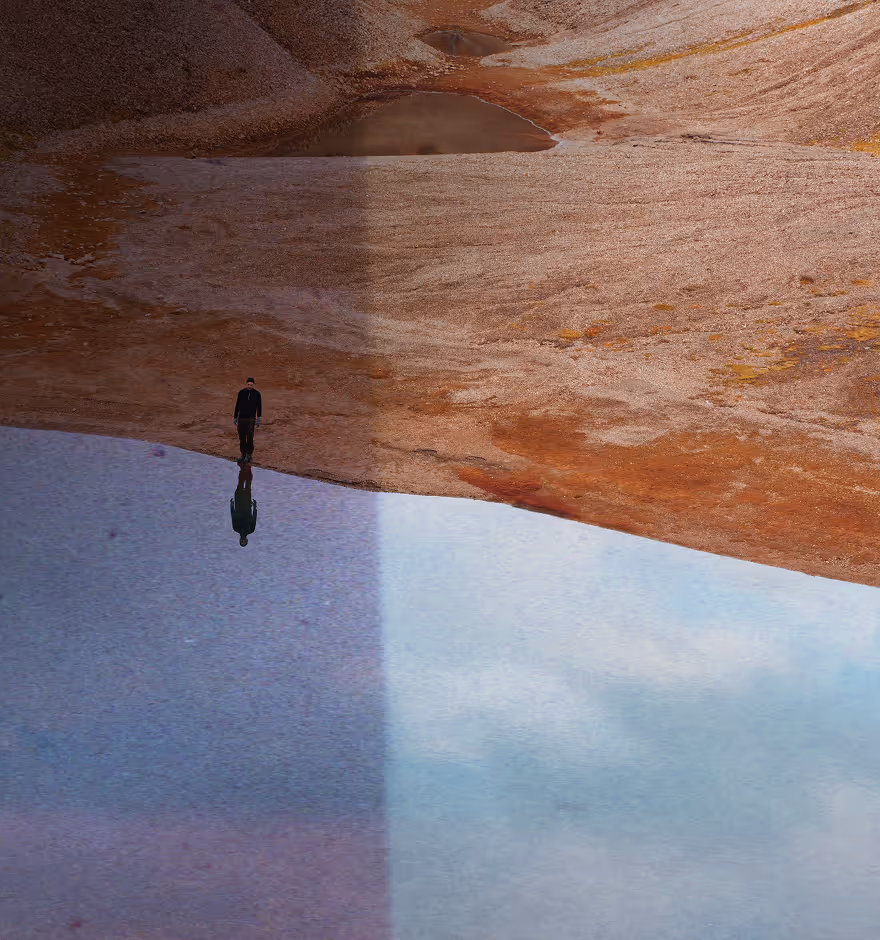 Film filter on a person standing alone on the edge of a calm body of water reflecting their silhouette and the surrounding barren, reddish terrain.
