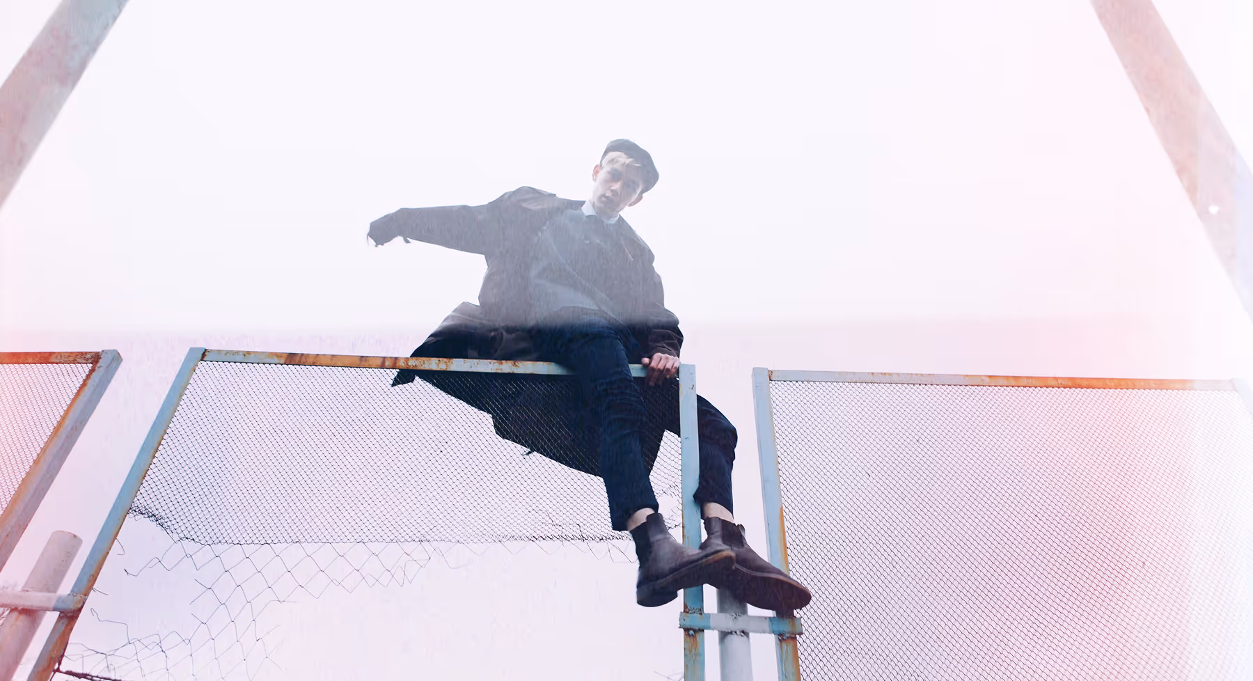 Cinematic effects on image of a young man in dark clothing and cap sitting atop a rusty metal fence against a pale sky.