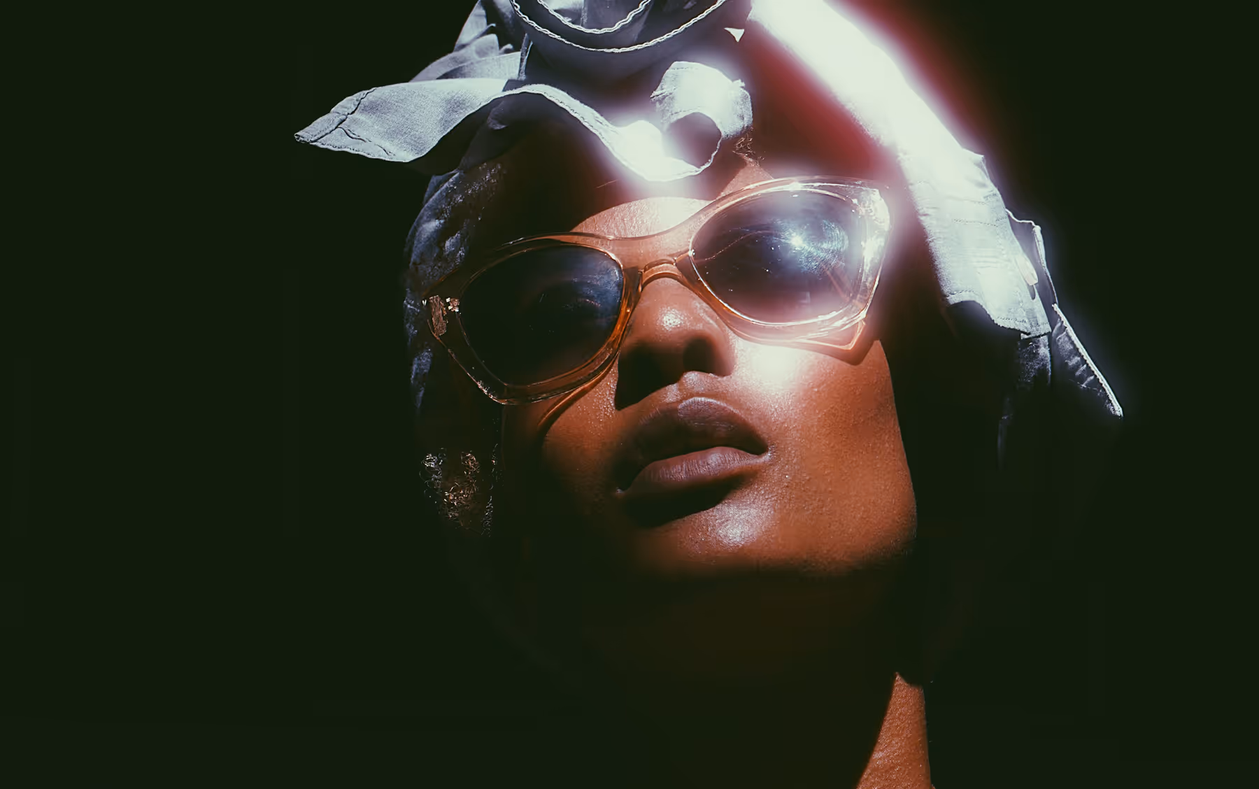 Cinematic close-up of a person wearing cat-eye sunglasses and a head wrap, illuminated by a bright light against a dark background.