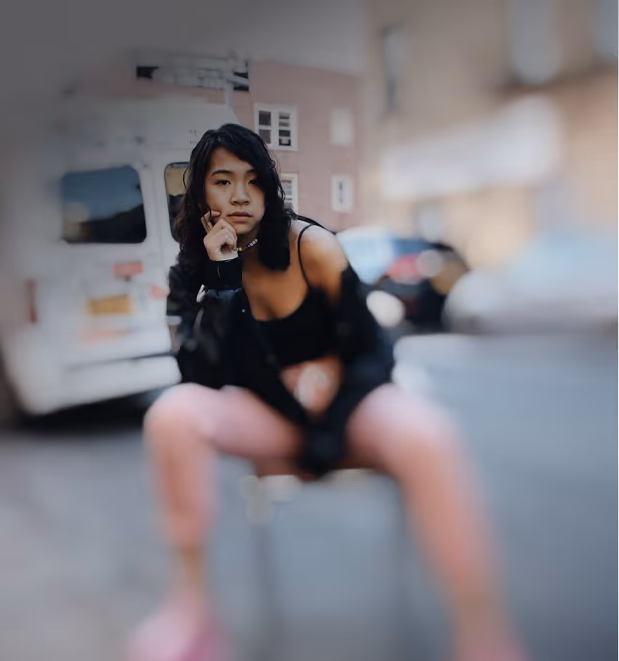 Young woman with dark hair sitting on a chair outdoors, resting her chin on her hand, wearing a black top and pink pants with a blurred city street background.