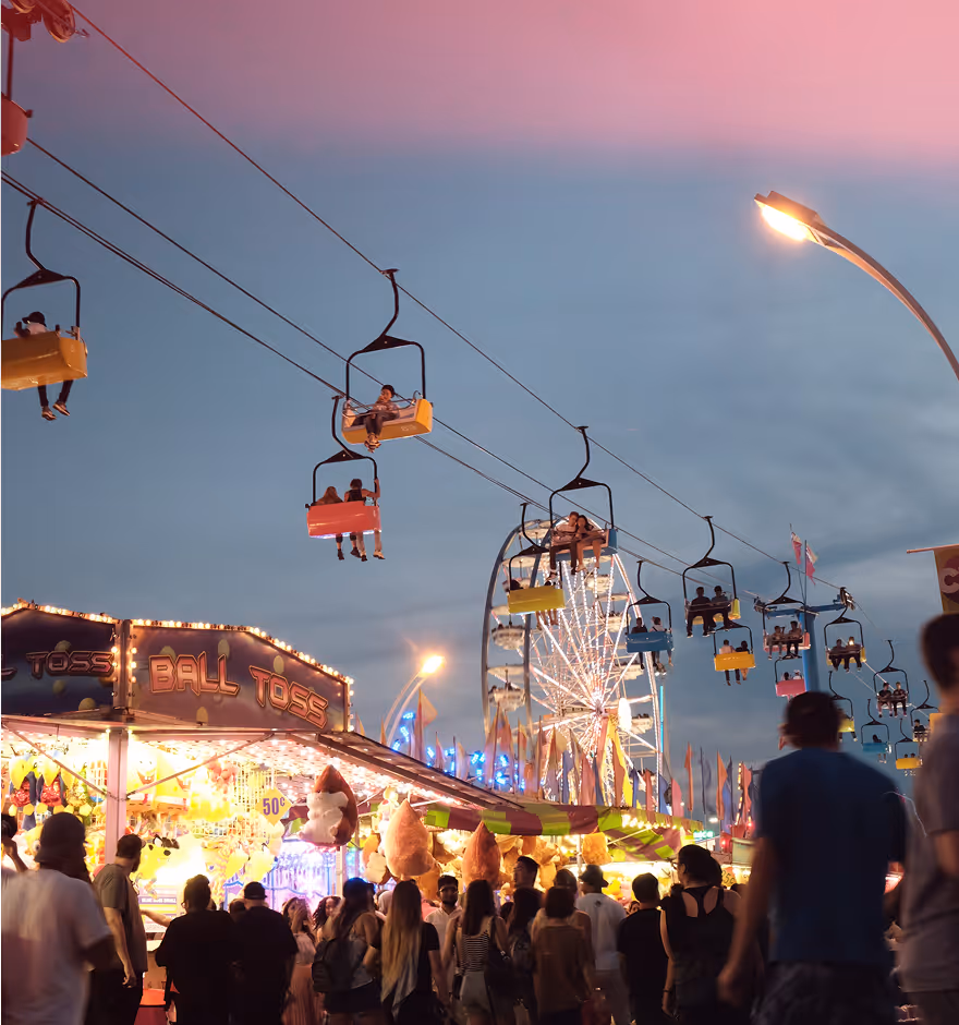 Light leak overlay on an evening carnival scene with people, colorful game stalls, a Ferris wheel, and sky chairs carrying riders overhead.
