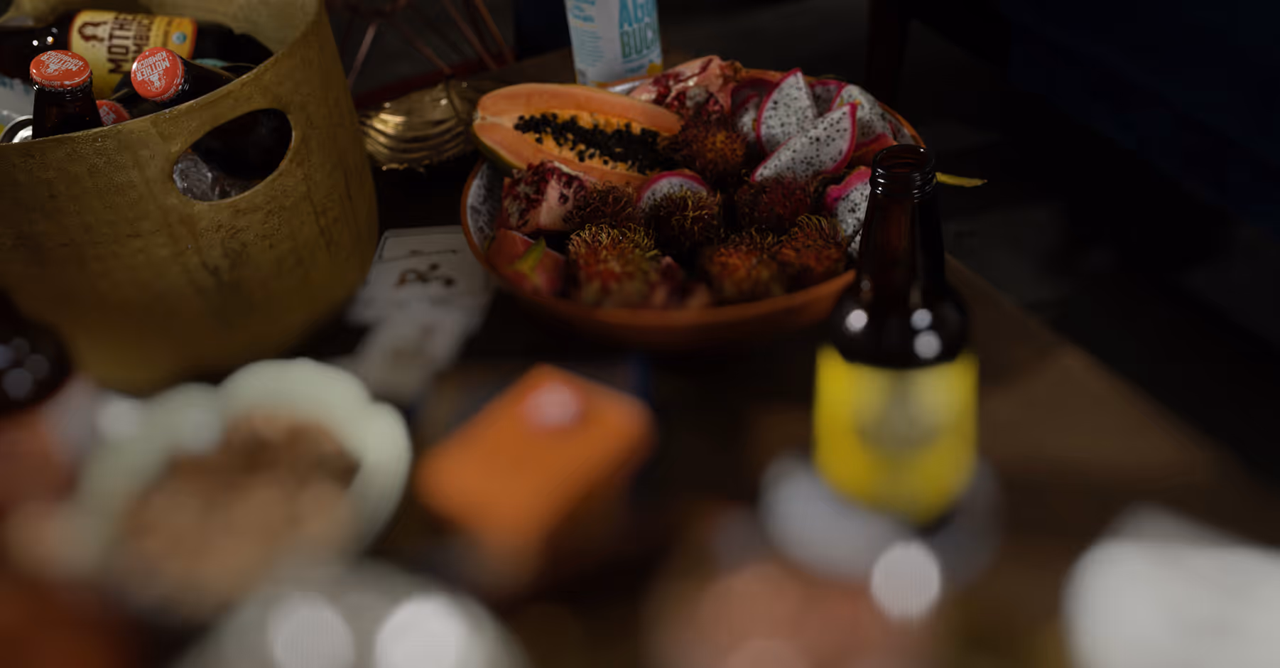 Tilt shift blur on bowl of tropical fruits including papaya, dragon fruit, pomegranate, and rambutan on a table next to a bucket of bottled drinks.