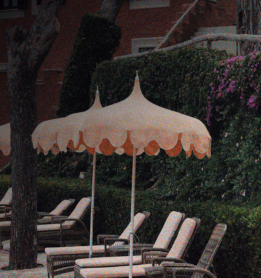 Grainy photo of wicker lounge chairs with cushions under scalloped-edge umbrellas beside a green hedge with flowers.