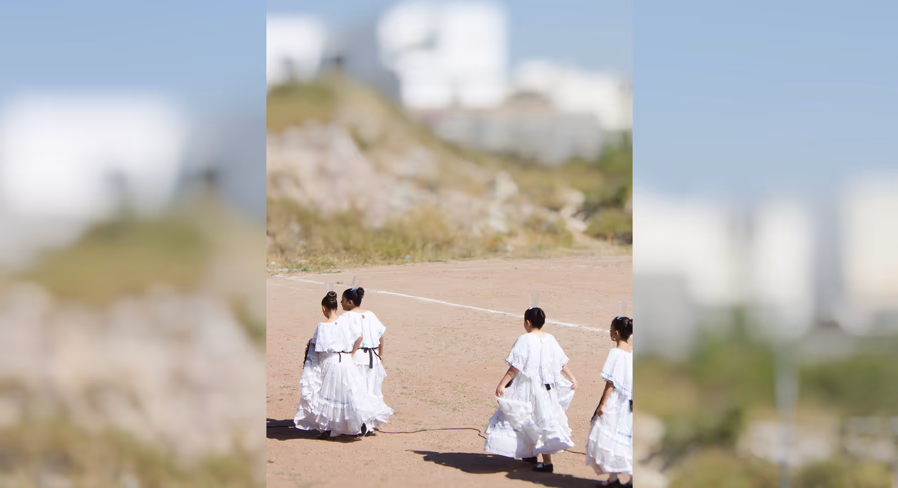 Tilt shift effect on image of four people in white dresses walking on a dirt field with a hilly background.